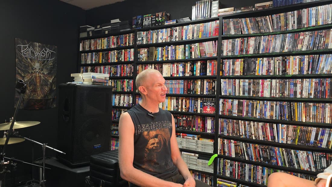 Cameron Novack seated in front of the store's wall of DVDs and VHS tapes during a filmed interview.