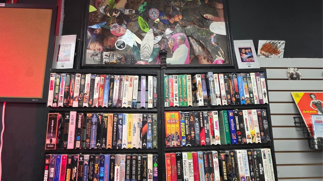 A wall of VHS tapes on black shelving with collage artwork mounted above.